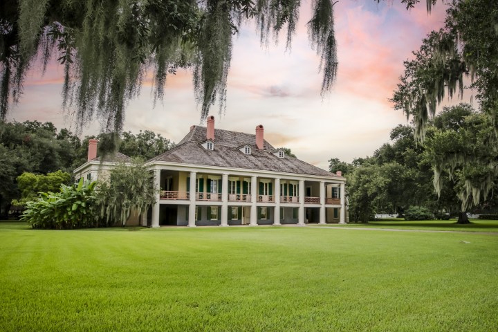 Historic mansion with columns and large lawn, surrounded by trees and hanging moss, at sunset.