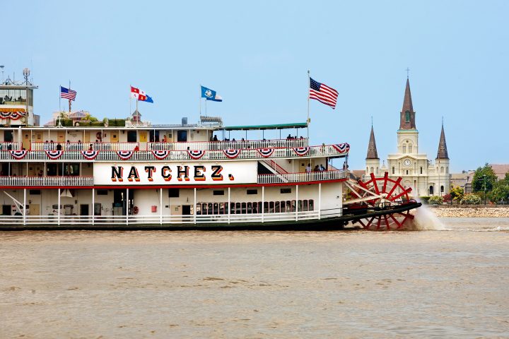 Steamboat Natchez with red paddlewheel in river, St. Louis Cathedral in background, blue sky.