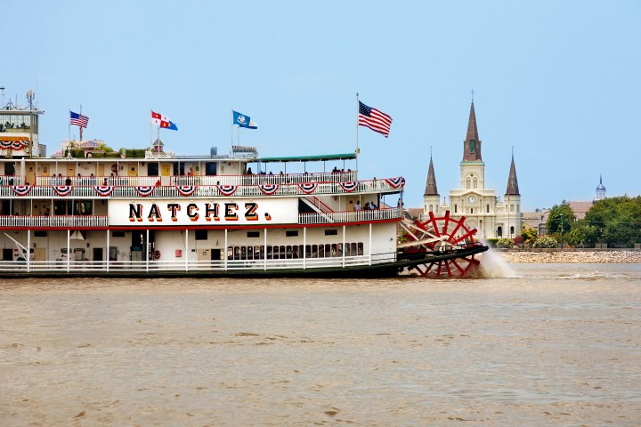 Steamboat Natchez with red paddlewheel in river, St. Louis Cathedral in background, blue sky.