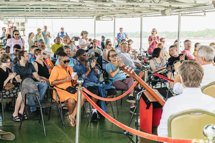 Audience on a boat watching a performance, people clapping, diverse crowd, sunny day.