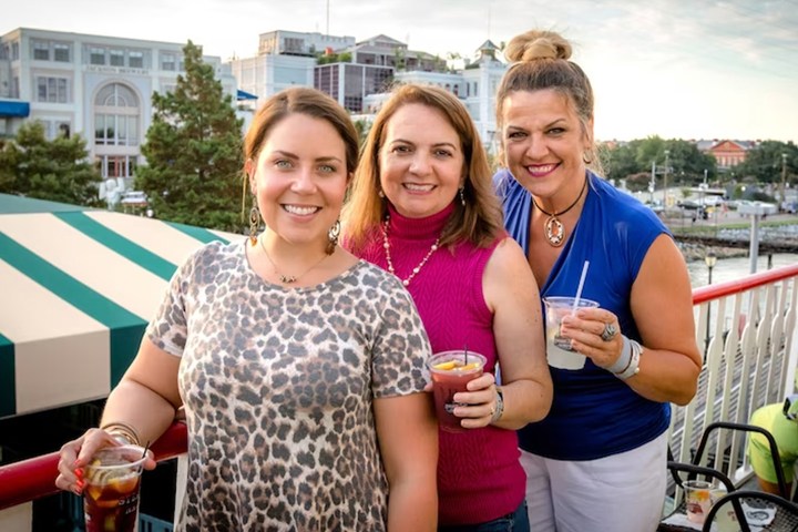 Three women smiling on a balcony holding drinks, with buildings and trees in the background.