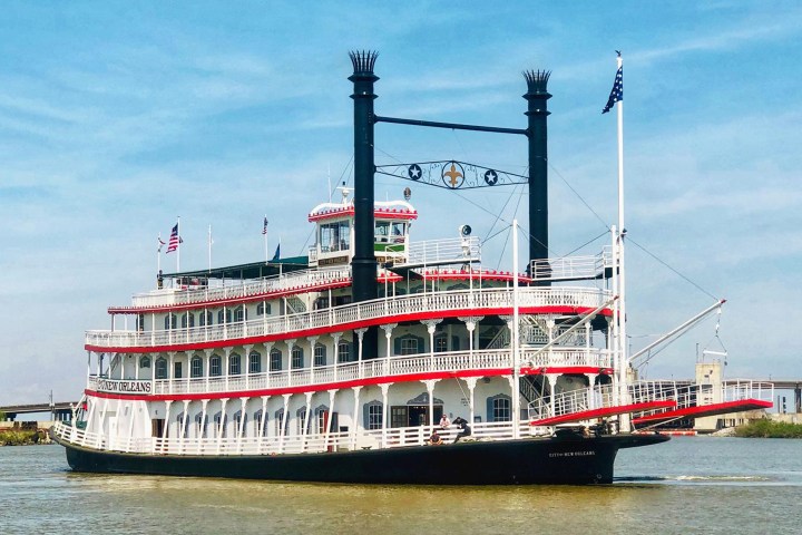 Paddle steamboat with red, white, and black trim on river, flags flying, under blue sky.
