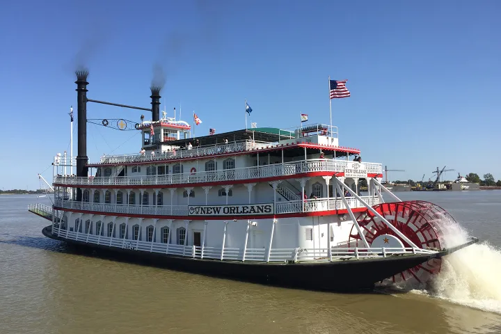Paddle steamer with 'New Orleans' sign sailing on river under clear blue sky.