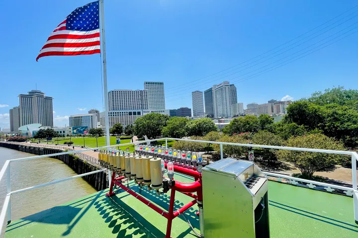 Riverboat calliope with American flag, city skyline, and trees on a sunny day.