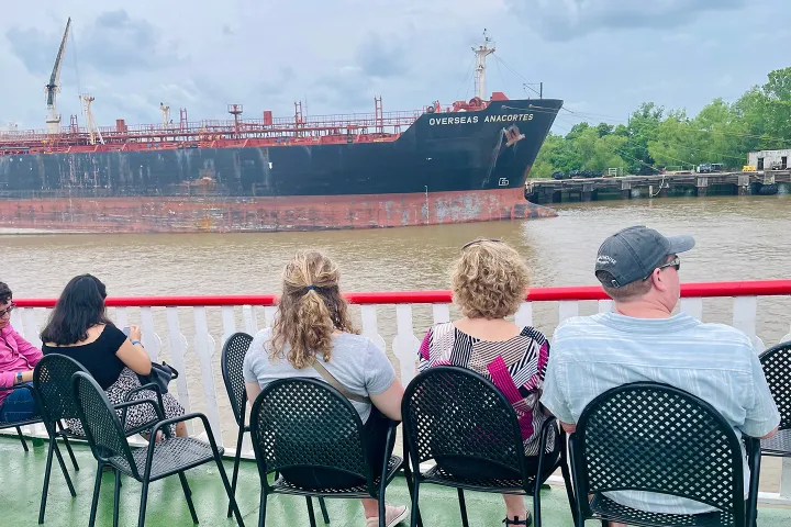 People seated on a boat deck watch a large cargo ship pass by on a river.