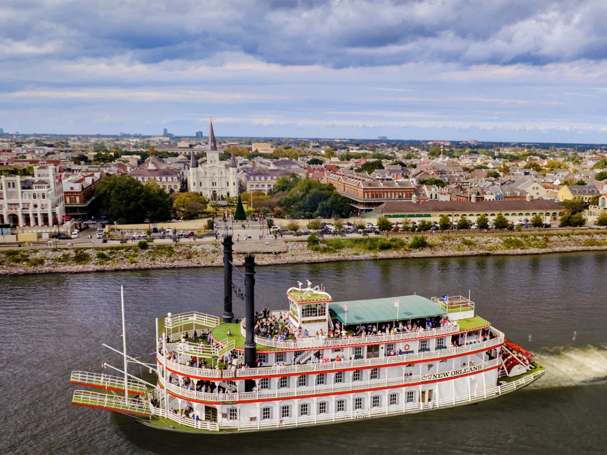 Riverboat cruising on a river with a cityscape in the background under cloudy skies.