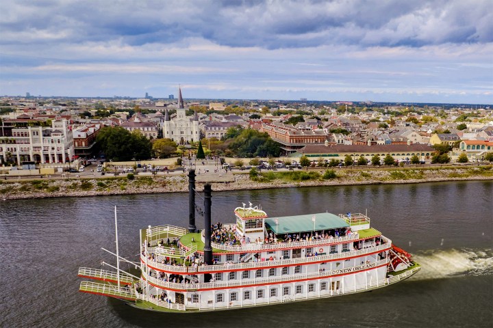 Riverboat cruising on a river with a cityscape in the background under cloudy skies.