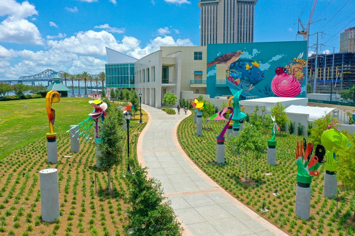 Colorful sculptures line a curved path near a modern building and large mural, under a blue sky.