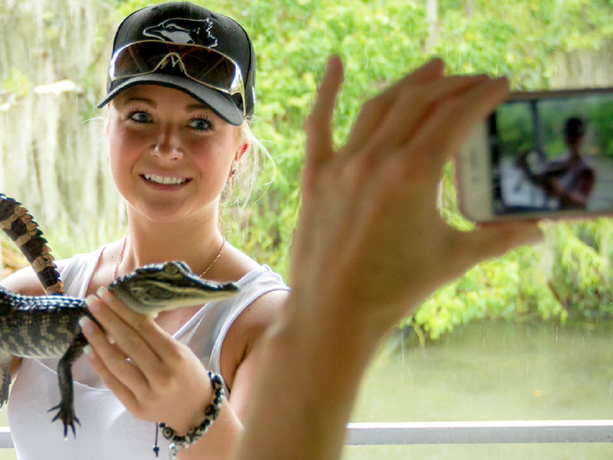 Person holding a small alligator while being photographed outdoors.