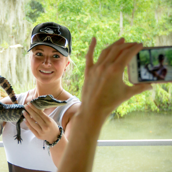 Person holding a small alligator while being photographed outdoors.