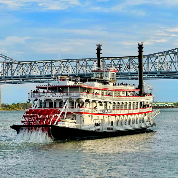 Paddle steamboat on river with bridge and skyline in background.