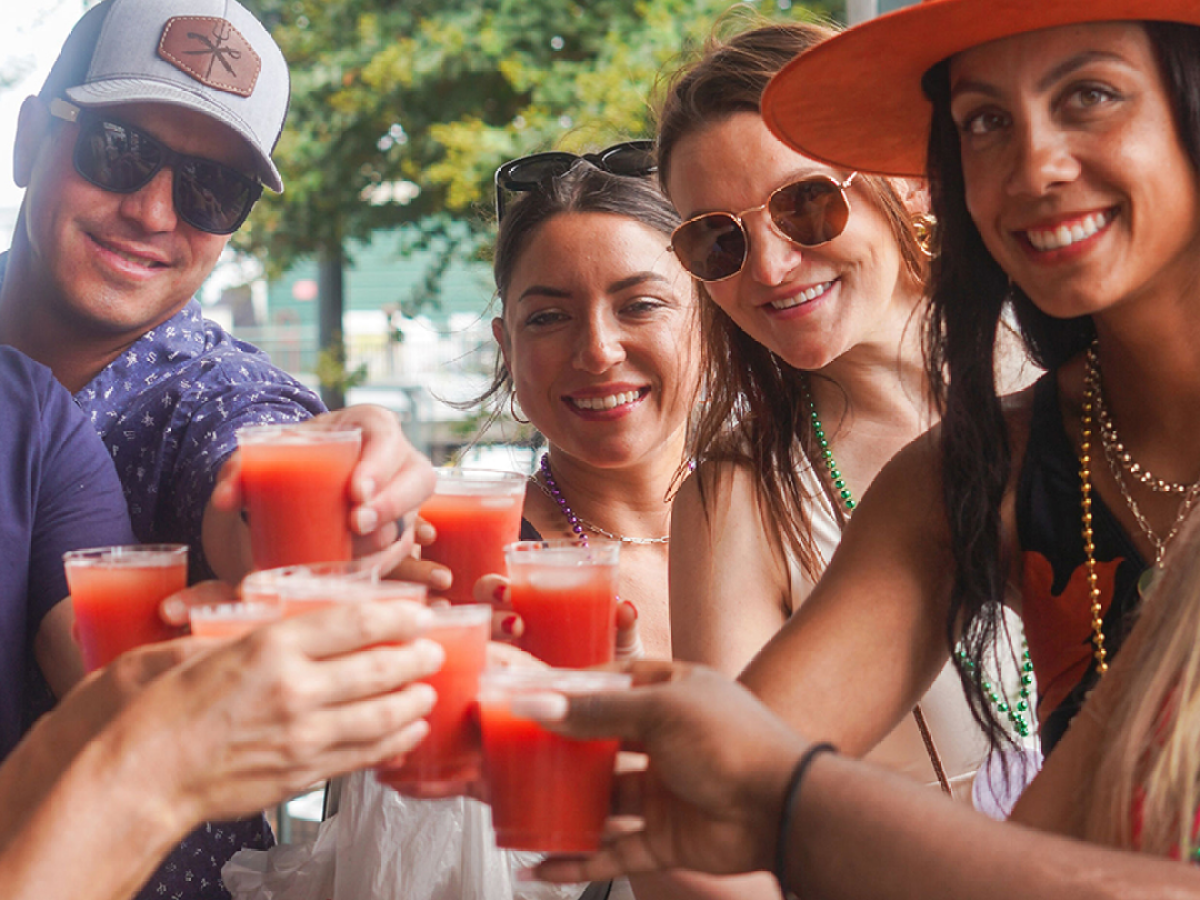 Group of people outdoors toasting with red drinks, wearing sunglasses and hats.