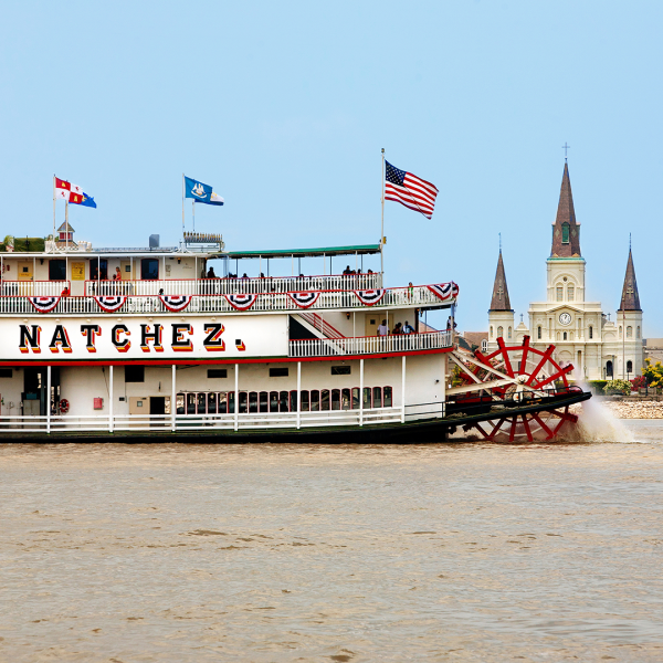 Paddle steamboat on river with church and flags in background.