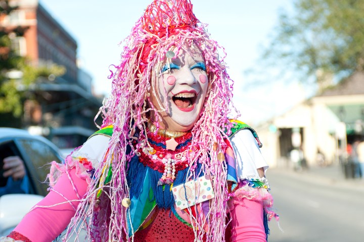 Person in colorful clown costume with pink hair smiling on a street.