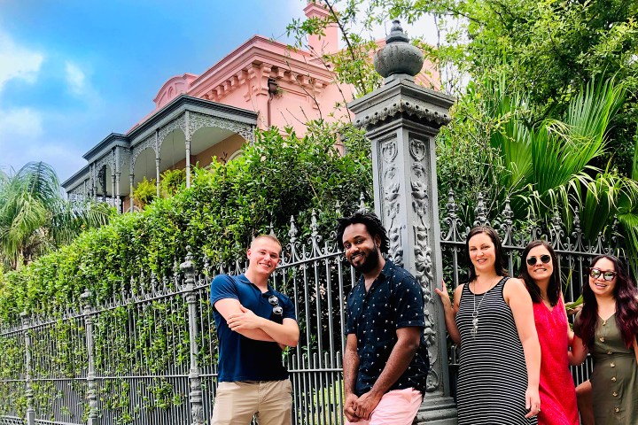 Group of people posing in front of a pink house with ornate fence and lush greenery.