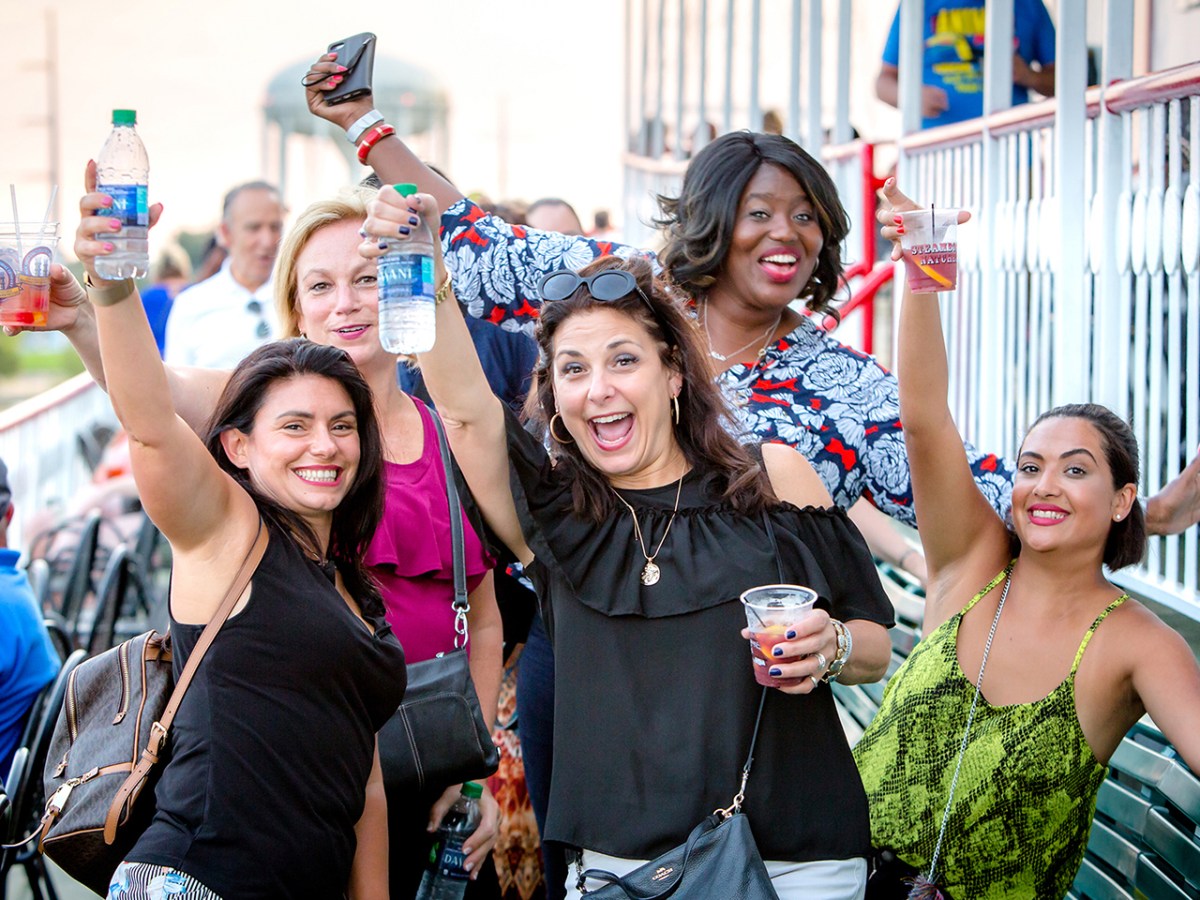 Group of five smiling women holding drinks and posing on a deck.