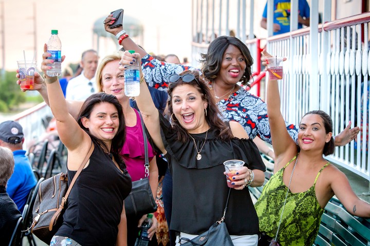 Group of five smiling women holding drinks and posing on a deck.