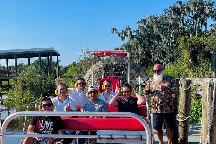 Group of seven people on a red airboat by a dock with trees in the background.