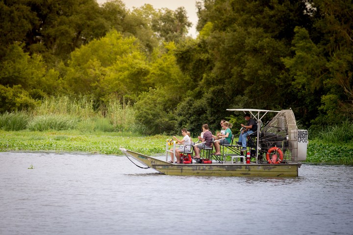 People riding an airboat on a river surrounded by trees.