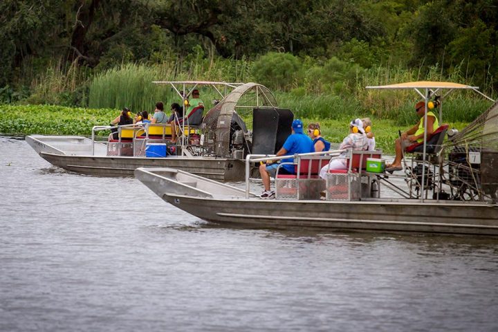 Two airboats with people on a river, surrounded by trees and greenery.
