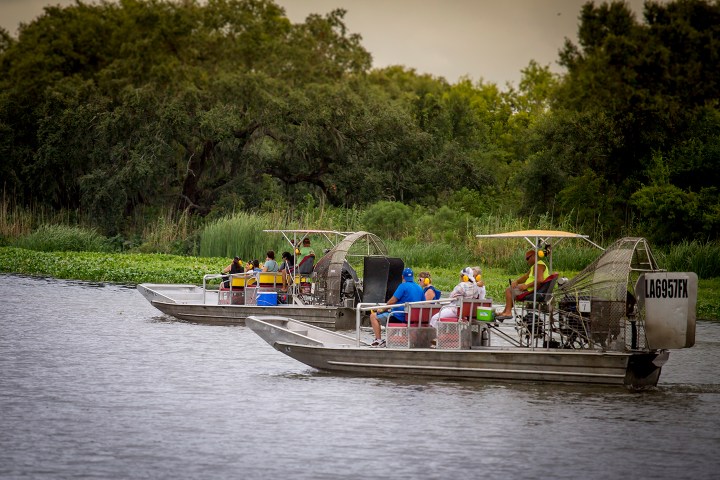 Two airboats with people on a river, surrounded by trees and greenery.