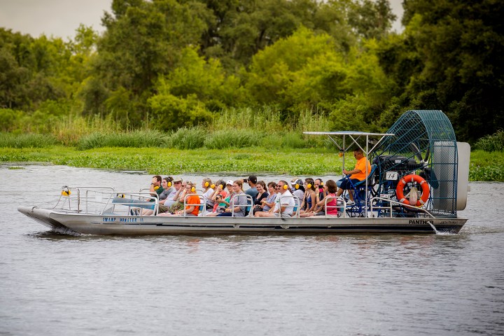 Group of people on an airboat in a river with green trees in background.