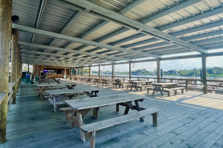 Covered outdoor seating area with wooden picnic tables by a waterfront.